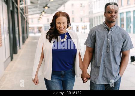 Fröhliches Paar schlendert Hand in Hand durch eine pulsierende Stadt und lacht unter der Sonne. Stockfoto