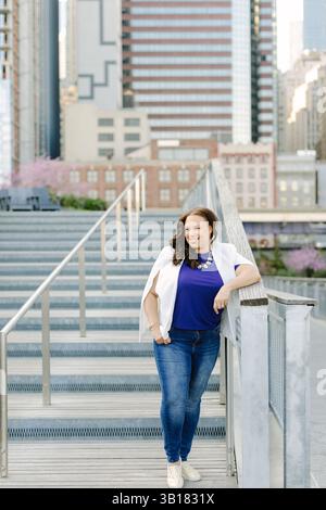 Frau in stilvoller Freizeitkleidung posiert bei Tageslicht auf einer städtischen Treppe mit der Skyline der Stadt im Hintergrund Stockfoto