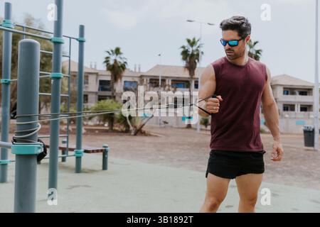 Ein Mann in einem roten Tanktop und schwarzen Shorts hält ein Widerstandsband. Die Szene spielt in einem Park mit ein paar Bänken und ein paar Palmen. Das mA Stockfoto