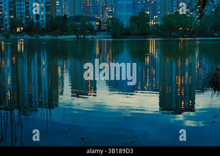 Eine ruhige abendliche Szene eines Sees, der Lichter der Stadt, Gebäude und Wasser reflektiert und in der Abenddämmerung einen Spiegeleffekt erzeugt. Schilf säumen die Wasserränder. Stockfoto