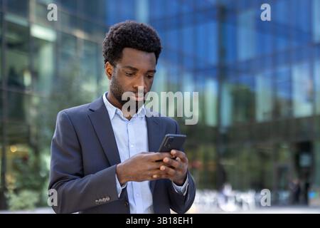 Seriöser, konzentrierter Geschäftsmann, der die App am Telefon nutzt und E-Mails liest. Afroamerikaner im Business-Anzug, der mit Smartphone in den Händen durch die Stadt geht. Stockfoto