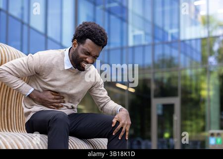 Mann mit Bauchschmerzen, Geschäftsmann, der auf einer Bank vor einem Bürogebäude sitzt und Hände an der Seite hält, Patient hat Entzündungen und schlechte Verdauung. Stockfoto