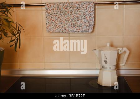 Kiew, Ukraine. 25. April 2025 Bialetti Moka-Topf, bereit zum Kaffeebrühen, auf einem schwarzen Kochfeld in einer gemütlichen Küche, neben einem Handtuch mit Blumenmuster Stockfoto
