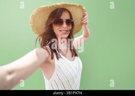 Eine 30- 35-jährige Touristenreisefrau mit breiter Krempe macht ein Selfie vor einem hellgrünen Hintergrundporträt isoliert. Das Konzept von Stockfoto