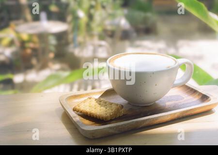 Frühstück im Freien - Eine Tasse warmen Cappuccino-Kaffee in einer Tasse aus weißem Ton mit Keksen auf einem Holztablett in einer üppigen Umgebung Stockfoto
