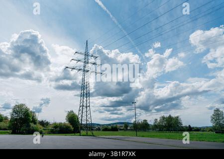 Stromleitungen und Türme erstrecken sich über eine ländliche Landschaft unter einem blauen Himmel mit flauschigen Wolken. Ein einsamer Radfahrer fährt auf der Straße unter der Weite Stockfoto