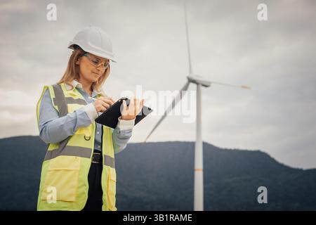 Eine Ingenieurin in einem Schutzhelm und einer gelben Weste inspiziert Daten auf einem Tablet an einem Windkraftstandort, umgeben von hohen Windturbinen, die Fortschritte bei der erneuerbaren Energie darstellen Stockfoto