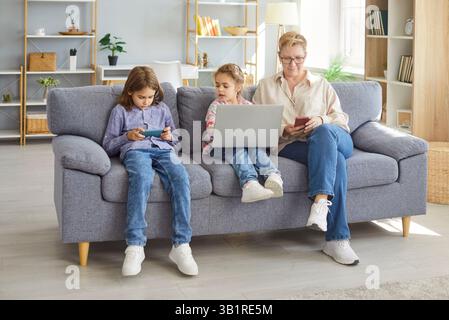 Ältere Großmutter mit Enkelkindern, die zusammen mit Laptop und Smartphones auf dem Sofa sitzen Stockfoto