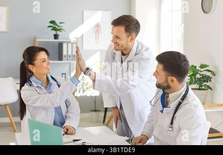 Drei lächelnde Ärzte feiern erfolgreiche Arbeit und geben hohe fünf an Kollegen in der medizinischen Praxis. Stockfoto