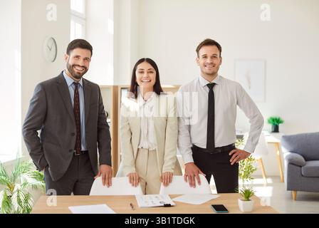 Drei Profis stehen am Schreibtisch, lächeln in die Kamera und zeigen einen positiven Teamgeist. Stockfoto