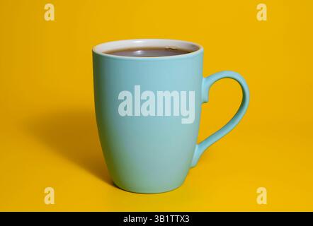 Ein hoher, mattblauer Keramikbecher mit Kaffeezubereiter auf einem leuchtend gelben Hintergrund mit sanfter Beleuchtung und einer klaren, minimalistischen Einrichtung. Stockfoto