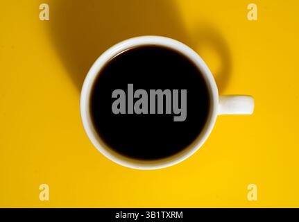 Überkopfaufnahme einer weißen Keramikkkaffeetasse mit schwarzem Kaffee auf einem leuchtend gelben Hintergrund mit sichtbarem Schatten. Stockfoto