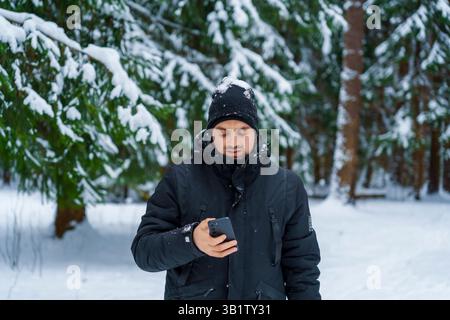 Junger Mann in schwarzem Wintermantel und Mütze, der in einem schneebedeckten Wald steht und das Smartphone mit Schnee auf dem Hut ansieht. Stockfoto