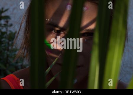 Detaillierte Nahaufnahme der Augen einer schönen Frau mit kreativem Neon-Make-up, das ihren Teil des Gesichts durch Palmblätter zeigt. Stockfoto