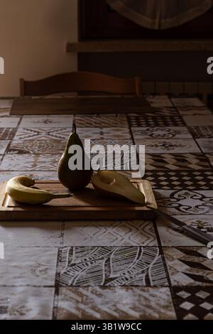 Moody Stillleben mit Birne und Banane auf einem Holzbrett. Das dramatische seitliche Licht schafft eine rustikale, gemütliche Atmosphäre in einer dunklen Küche. Stockfoto