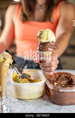 Nahaufnahme eines Eiskegels mit zwei Kugeln verschiedener Geschmacksrichtungen, gehalten von einer jungen Frau in einer Küche. Desserts, gastronomisches Branding oder Werbeaktion Stockfoto