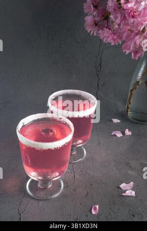 Zwei kleine Gläser, gefüllt mit einem rosafarbenen Getränk, mit Zucker umrandet, auf einer dunklen Oberfläche mit herabfallenden rosa Kirschblüten Stockfoto