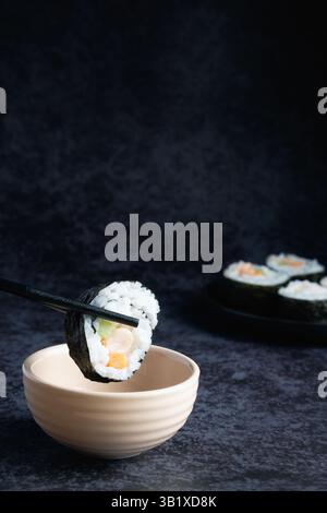 Eine Nahaufnahme zeigt Stäbchen, die ein Stück Sushi-Rolle mit sichtbarem Reis, Algen und Füllung über einer Schüssel mit Sauce halten. Anderer Sushi-Kuchen Stockfoto