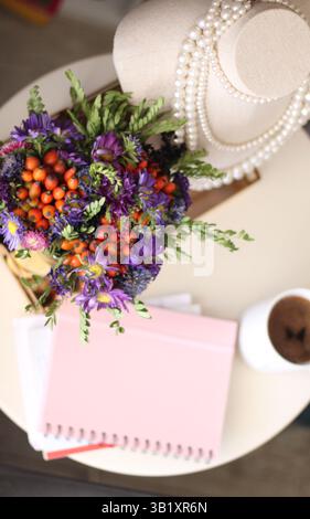 Blick von oben auf einen gemütlichen, femininen Arbeitsplatz mit Blumenstrauß, Perlenkette, Kaffeetasse und Notizbuch in Pastellfarben. Sanftes Morgenlicht mit Kaffee Stockfoto