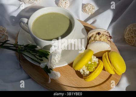 Süßes gelbes Macaron Dessert Tasse grüner Matcha Tee Latte auf Pflanzenbasis in gemütlicher Morgenliegenatmosphäre. Kreative Urlaubsidee mit romantischem Date. Pflanzliche präbiotische Faser Superfood Getränk Antioxidans-reiche Immunität-Boosting Kaffee Alternativen Getränk Stockfoto