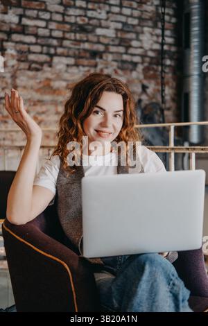 Eine junge Frau mit lockigen roten Haaren sitzt mit ihrem Laptop in einem Café und schaut selbstbewusst in die Kamera. Selbstbewusste Frau, moderne Unternehmerin, Startup Stockfoto
