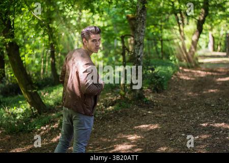 Ein junger Mann in lässiger Kleidung schlendert über einen unbefestigten Pfad in einem üppig grünen Wald. Sonnenlicht zieht durch die Bäume und schafft eine ruhige Atmosphäre, während er über seine Schulter zurückblickt. Stockfoto