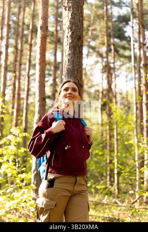Eine junge Frau steht in einem friedlichen Wald und genießt die Sehenswürdigkeiten und Klänge der Natur in lässiger Wanderkleidung und einem Rucksack. Stockfoto
