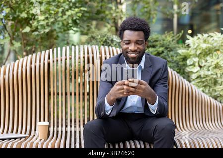 Ein lächelnder Geschäftsmann im Anzug überprüft sein Handy, während er draußen auf einer Holzbank sitzt. Eine Kaffeetasse und ein Laptop ruhen in der Nähe. Stockfoto
