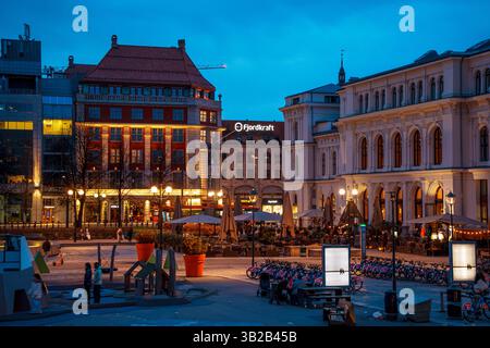 Oslo, Norwegen - 13. April 2024: Abendlicher Blick auf einen belebten Platz der Stadt Oslo mit beleuchteten Gebäuden und Cafés im Freien. Stockfoto