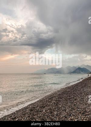 Dramatische Regenwolken über ruhigem Meer und Kiesstrand mit Bergen am Horizont Stockfoto