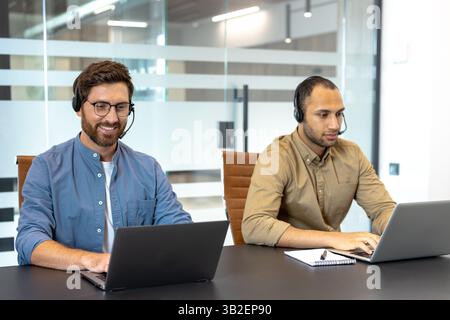 Zwei Kundendienstmitarbeiter mit Headsets arbeiten an Schreibtischen mit ihren Laptops in einer modernen Büroumgebung. Stockfoto