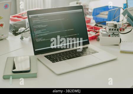 Mittelgroße Nahaufnahme eines silbernen Laptops mit Codes auf dem Bildschirm, Plastikkästen mit winzigen Details und elektronischem Gerät auf weißem Arbeitstisch Stockfoto