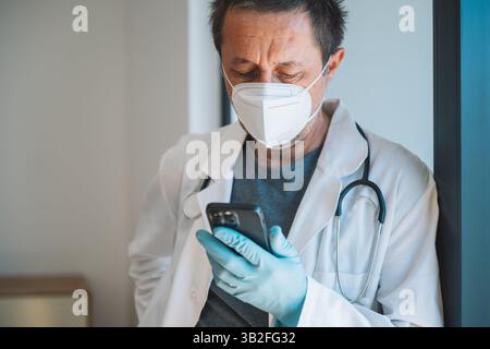 Müder erschöpfter männlicher Arzt trägt Schutzmaske mit Smartphone in Krankenhausklinikpraxis, selektiver Fokus Stockfoto