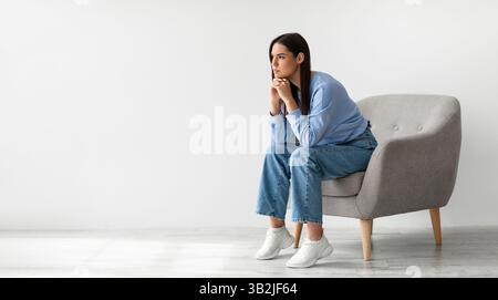 Porträt einer verärgerten jungen Dame, die im Sessel vor der weißen Studiowand sitzt, Bannerdesign mit freiem Platz Stockfoto