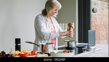 Laptop, Kochen und Seniorin in der Küche des modernen Hauses für gesunde oder Diät-Abendessen. Essen, Technologie und ältere Frau, die eine Mahlzeit zubereitet Stockfoto