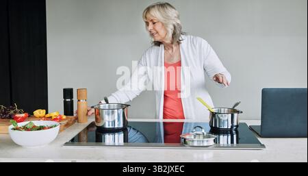 Laptop, Kochen und Seniorin in der Küche des modernen Hauses für gesunde oder Diät-Abendessen. Essen, Technologie und ältere Frau, die eine Mahlzeit zubereitet Stockfoto