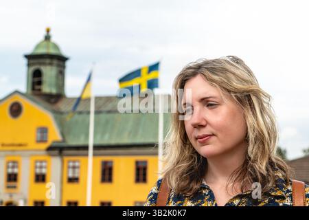 Eine Frau posiert nachdenklich mit den schwedischen und ukrainischen Fahnen, die an einem sonnigen Sommertag vor einem farbenfrohen Gebäude in Schweden steht. Stockfoto