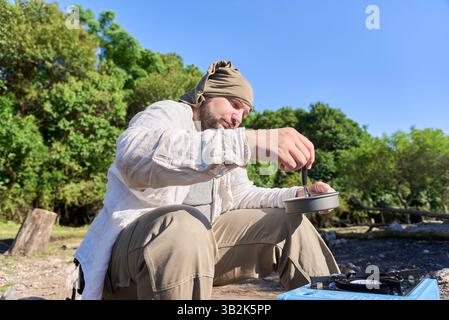 Hispanische Reisende kochen draußen auf einem hellblauen tragbaren Herd, während sie an einem sonnigen Morgen auf einem Naturcamping campen. Stockfoto
