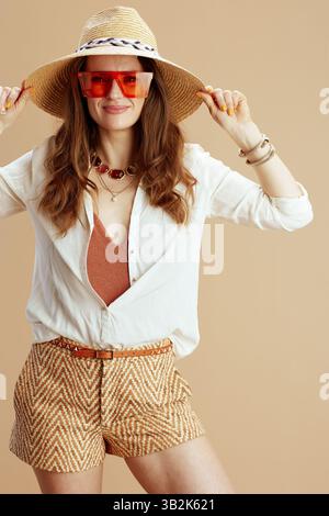 Strandurlaub. Lächelnde elegante 40 Jahre alte Hausfrau in weißer Bluse und Shorts isoliert auf Beige mit Strohhut und Sonnenbrille. Stockfoto