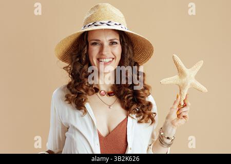 Strandurlaub. Lächelnde elegante 40-jährige Frau in weißer Bluse und Shorts auf beigefarbenem Hintergrund mit Seestern und Sommerhut. Stockfoto