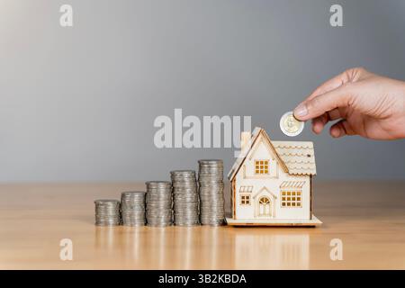 Der Mann wirft Münzen in ein Glas, mit Münzstapeln und einem hölzernen Hausmodell neben ihm. Betont das Konzept, Geld für zukünftige Einfamilienhäuser zu sparen Stockfoto