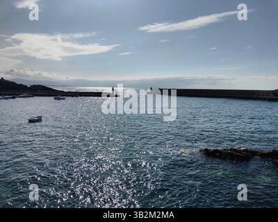 Glitzerndes Meereswasser in einem Fischerhafen mit Booten, Wellenbrechern, Leuchtturm und Hügeln an einem sonnigen Tag Stockfoto