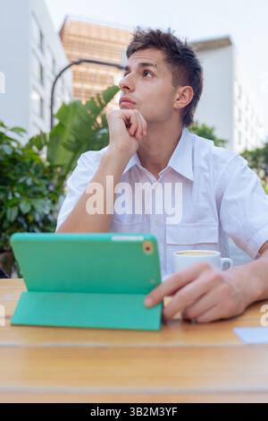 Junger Mann denkt, während er ein Tablet in einem Café im Freien in vertikaler urbaner Umgebung benutzt Stockfoto
