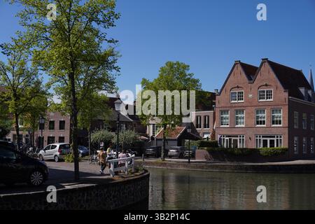 „Malerischer Kanal gesäumt von Booten, Autos und historischen Gebäuden unter üppigem Frühlingslaub in Hoorn, Niederlande. Stockfoto