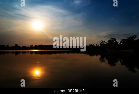 Ein ruhiger Sonnenuntergang über einem ruhigen See, während die Sonne auf der Wasseroberfläche reflektiert. Silhouetten von Bäumen säumen das Ufer und schaffen eine friedliche Atmosphäre. Stockfoto