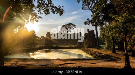 Ein ruhiger Blick auf alte Ruinen, umgeben von üppigem Grün, reflektiert in einem ruhigen Teich unter einem goldenen Sonnenuntergang. Die Szene fängt die Schönheit der Natur ein Stockfoto