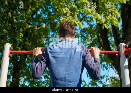 Sportlicher Mann, der Pull-ups auf der roten horizontalen Bar im Park macht. Athlet trainiert die Oberkörpermuskulatur im Outdoor-Gym. Person, die Krafttrainings durchführt. Co Stockfoto