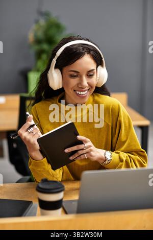 In einem lebhaften Büro macht sich eine junge Frau mit Kopfhörern gerne Notizen, während sie sich konzentriert. Stockfoto