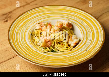 Ein wunderschön überzogener Garnelen-Spaghetti-aglio e olio wird auf einer gelb gestreiften Keramikschale auf einem Holztisch präsentiert. Die Teigwaren sind mit Olivenöl überzogen Stockfoto