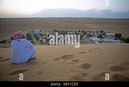 Beduinenmann in weißer Dschungadscha, der auf einer Sanddüne sitzt und ein Wüstencamp, Wahiba Sands Wüste oder Rimal Al Wahiba oder Sharqiyya Sands, Janub Ash-S, betrachtet Stockfoto